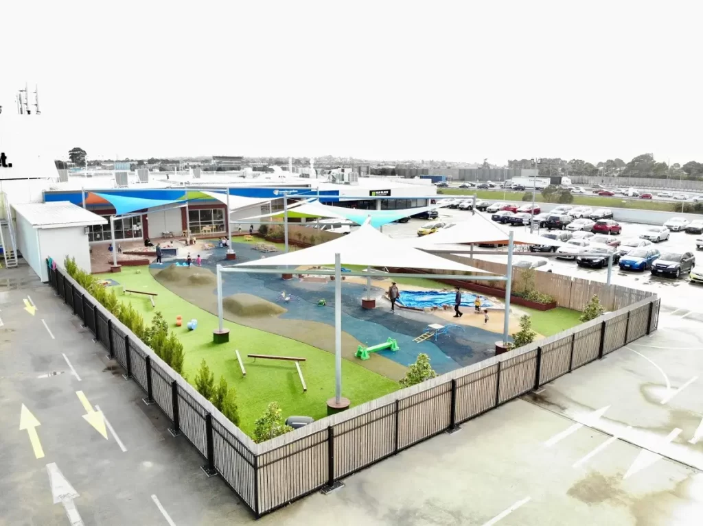 Elevated view of a modern early learning centre featuring a secure outdoor playground with shade sails, nature-inspired play areas, landscaped gardens, and dedicated learning spaces for children.