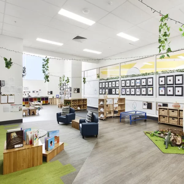 Bright early learning classroom featuring natural timber furniture, reading nooks, open play areas, learning stations, nature-inspired décor, and children's artwork displayed throughout the space.