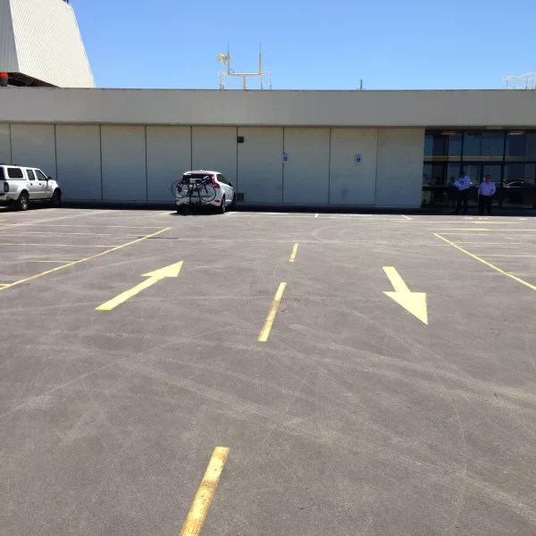 Large commercial building with an expansive asphalt car park featuring freshly painted directional arrows, parking bays, and a main building entrance under a clear blue sky.