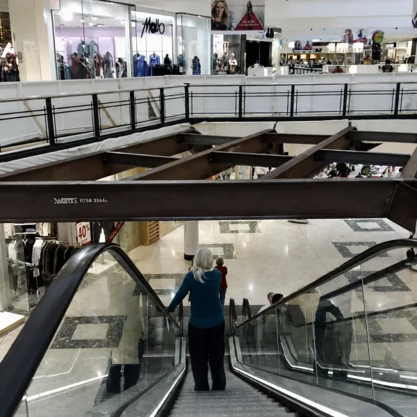 Shopping centre refurbishment in progress featuring exposed structural steel framing above an escalator, with retail stores remaining operational during the commercial fit-out.
