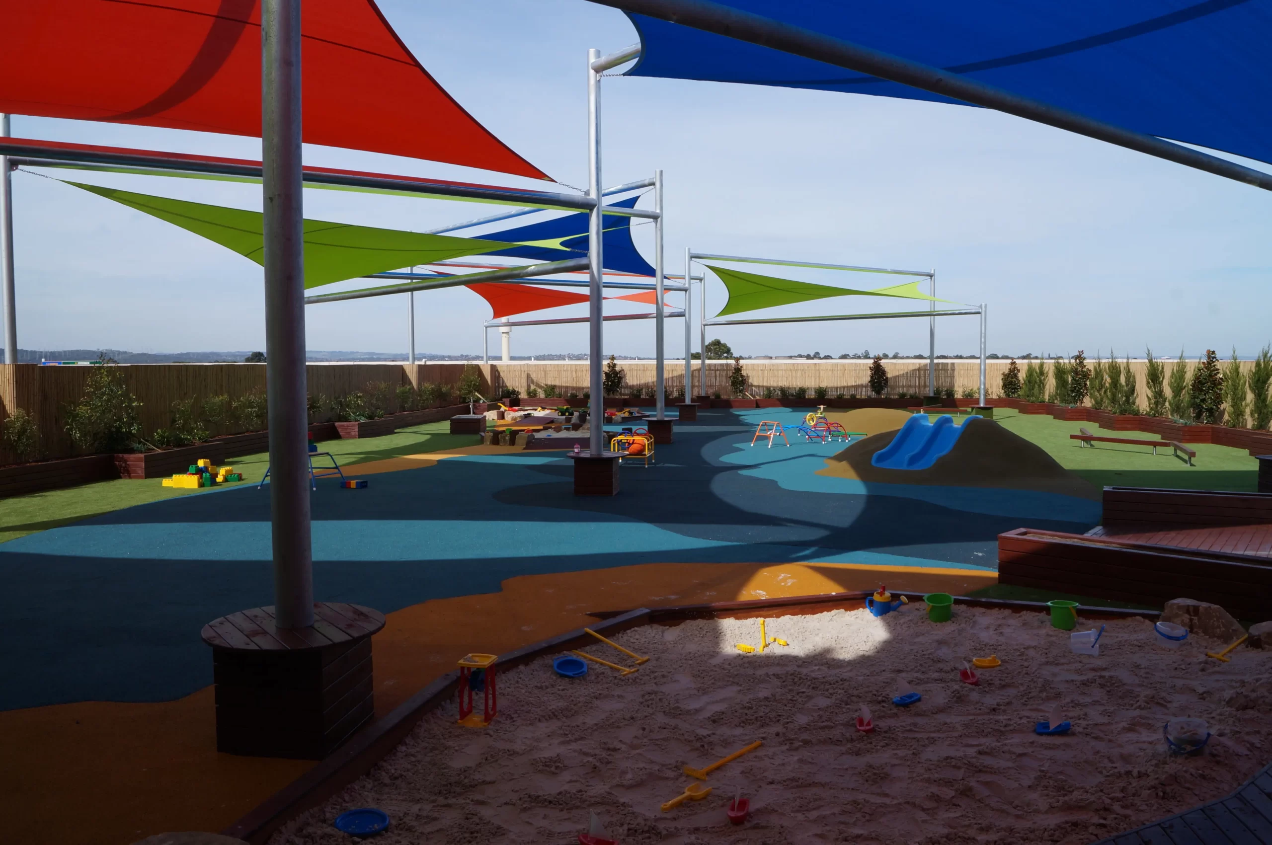 Purpose-built childcare outdoor playground featuring colourful shade sails, sandpit, climbing and play equipment, soft-fall surfaces, and landscaped play areas for early childhood learning.