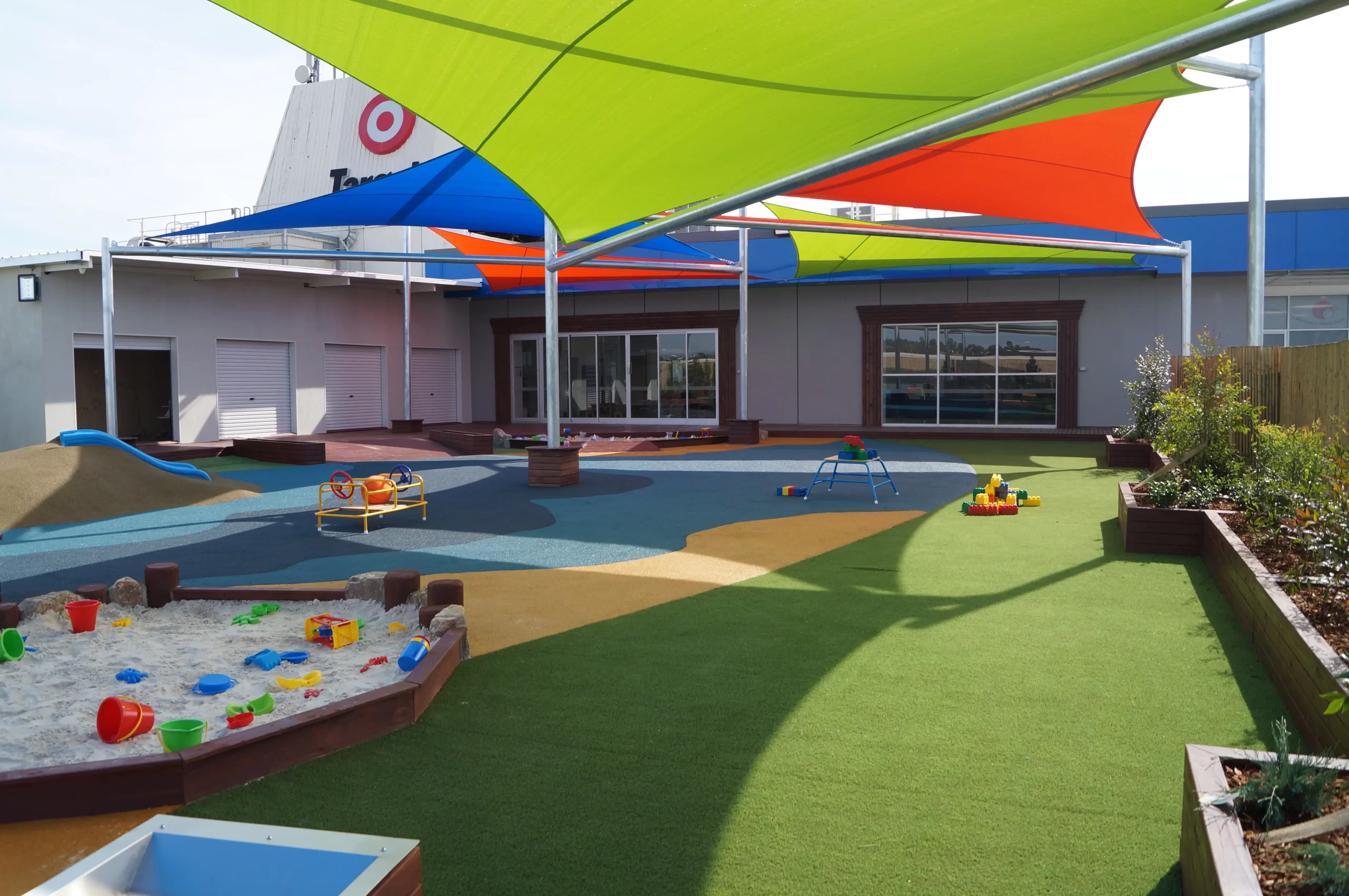 Modern childcare centre outdoor play area featuring colourful shade sails, artificial turf, sandpit, play equipment, and landscaped learning spaces designed for early childhood education.