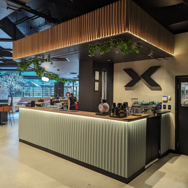Modern X-Golf reception and café bar featuring custom illuminated joinery, timber feature ceiling, integrated greenery, and contemporary commercial interior design.
