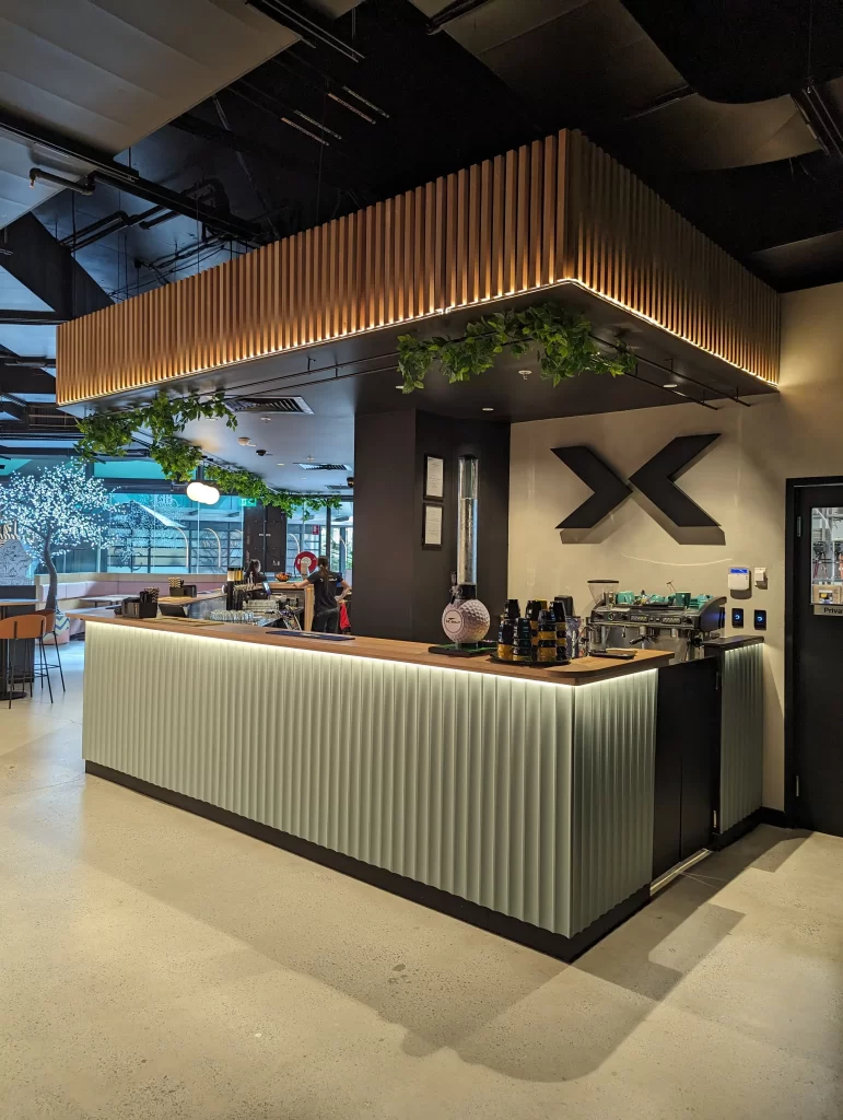 Modern X-Golf reception and café bar featuring custom illuminated joinery, timber feature ceiling, integrated greenery, and contemporary commercial interior design.