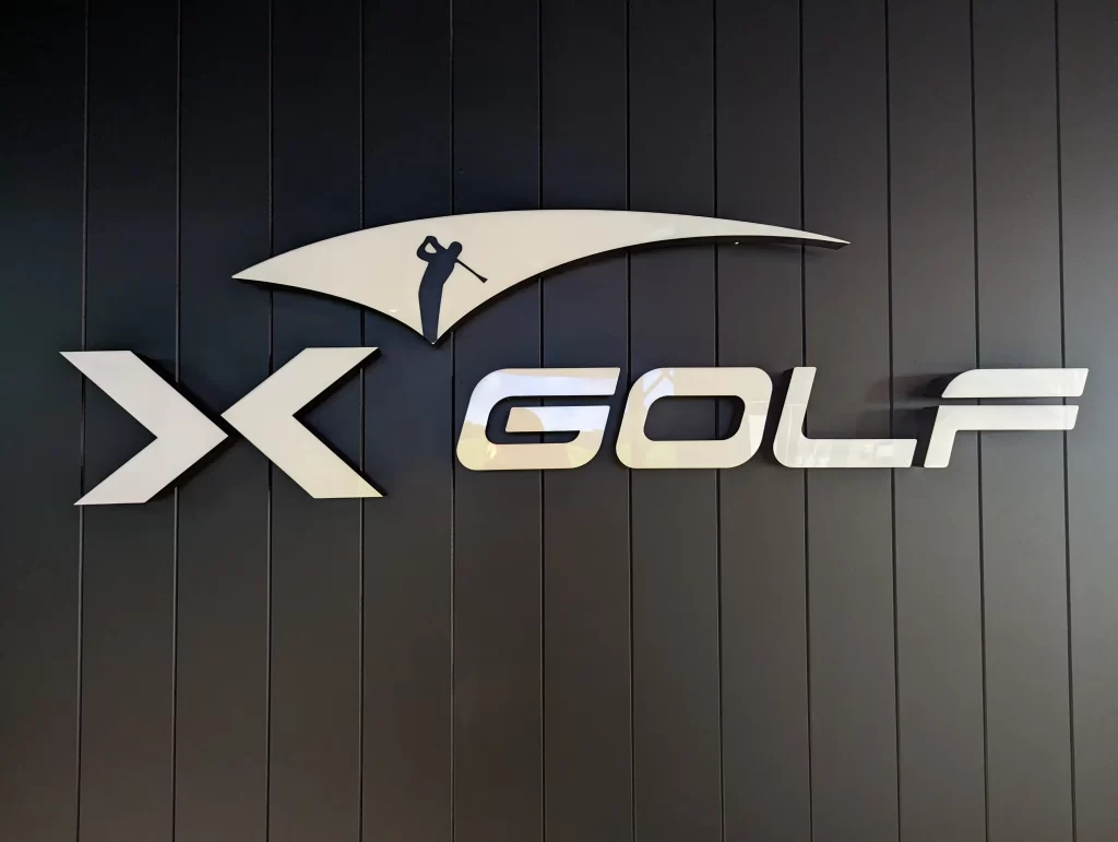 X-Golf illuminated logo mounted on a modern black feature wall inside a commercial indoor golf entertainment venue.