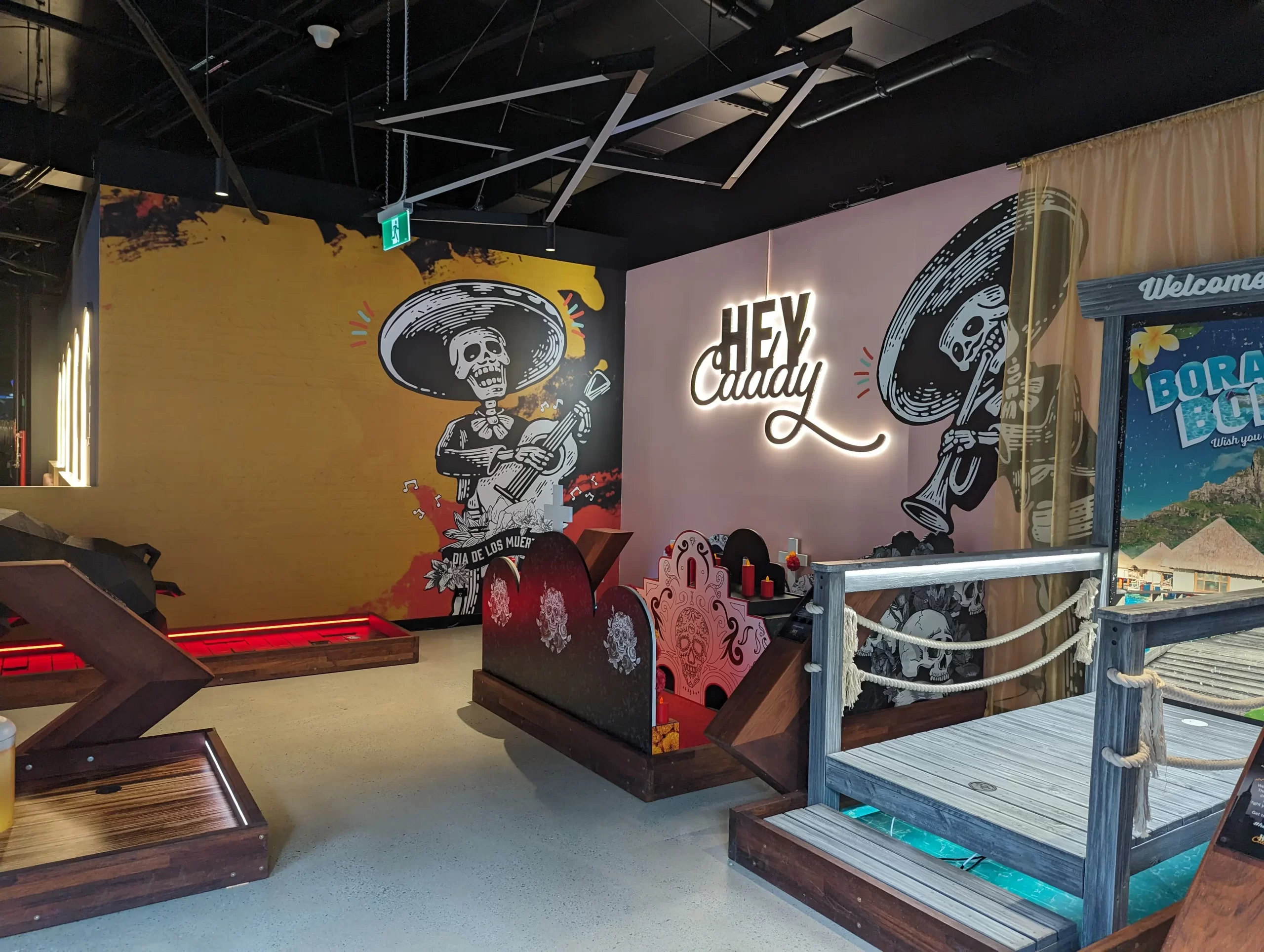 Themed indoor mini golf hole featuring vibrant Día de los Muertos murals, illuminated "Hey Caddy" signage, custom-built obstacles, and immersive entertainment venue design.