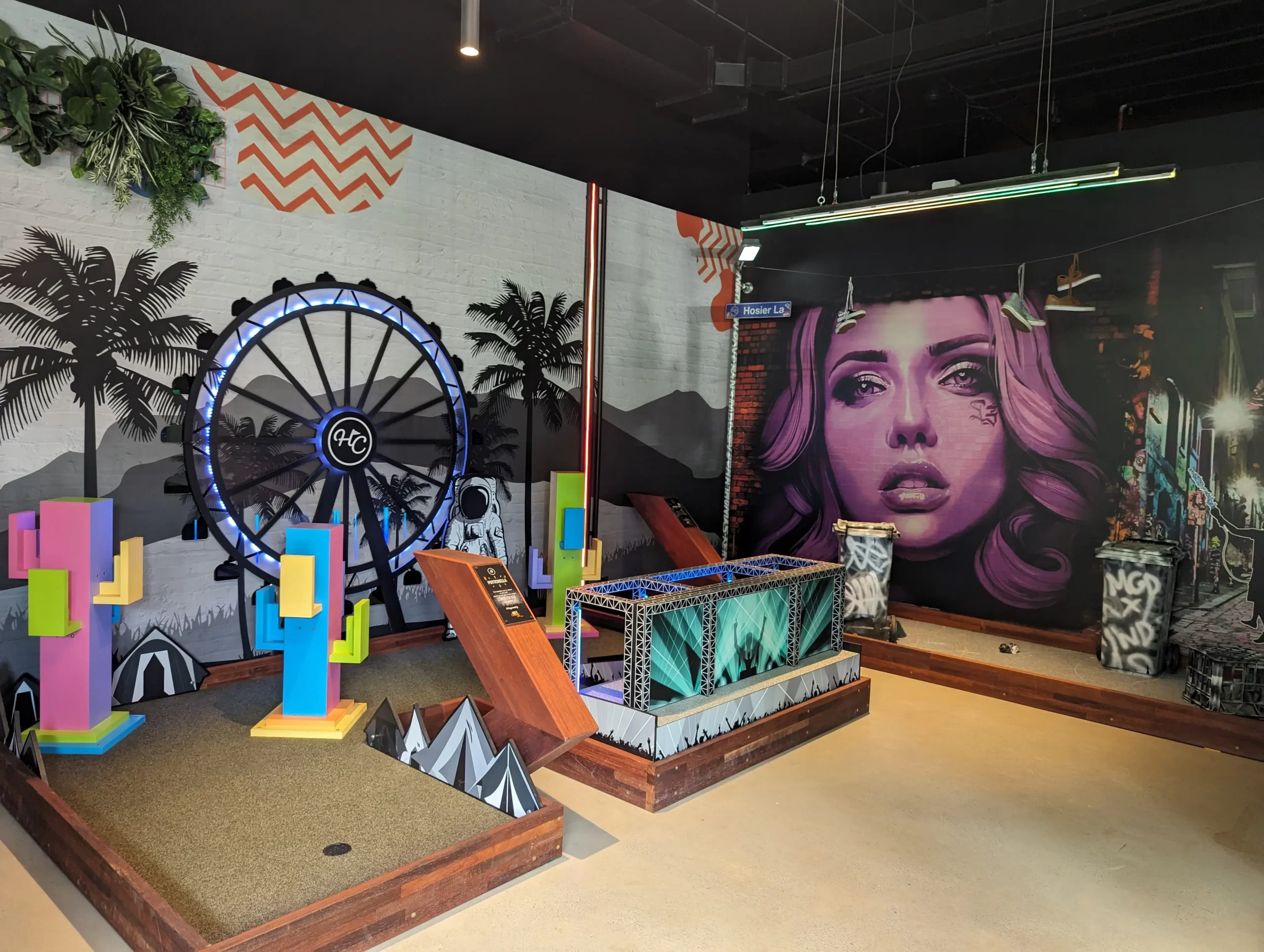 Immersive indoor mini golf course featuring a large illuminated water wheel, colourful custom obstacles, vibrant street art murals, and themed entertainment venue design.