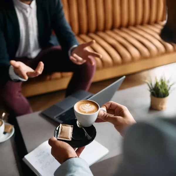 Business professionals meeting over coffee with a laptop and notebook, discussing commercial interior design concepts and project planning in a modern lounge setting.