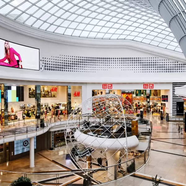 Multi-level shopping centre atrium with a large geometric installation beneath a curved glass skylight, surrounded by retail stores.