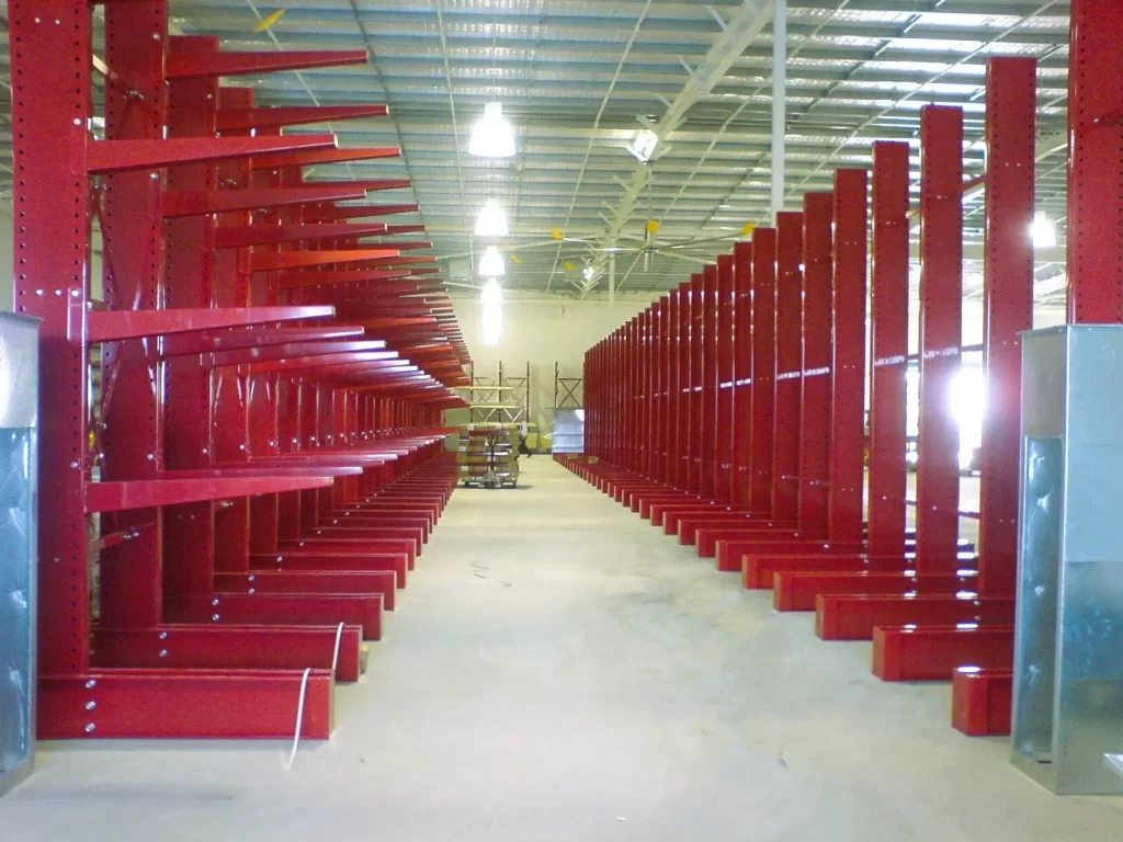 Wide warehouse aisle with red cantilever storage racks on both sides beneath a high industrial ceiling with overhead lighting.