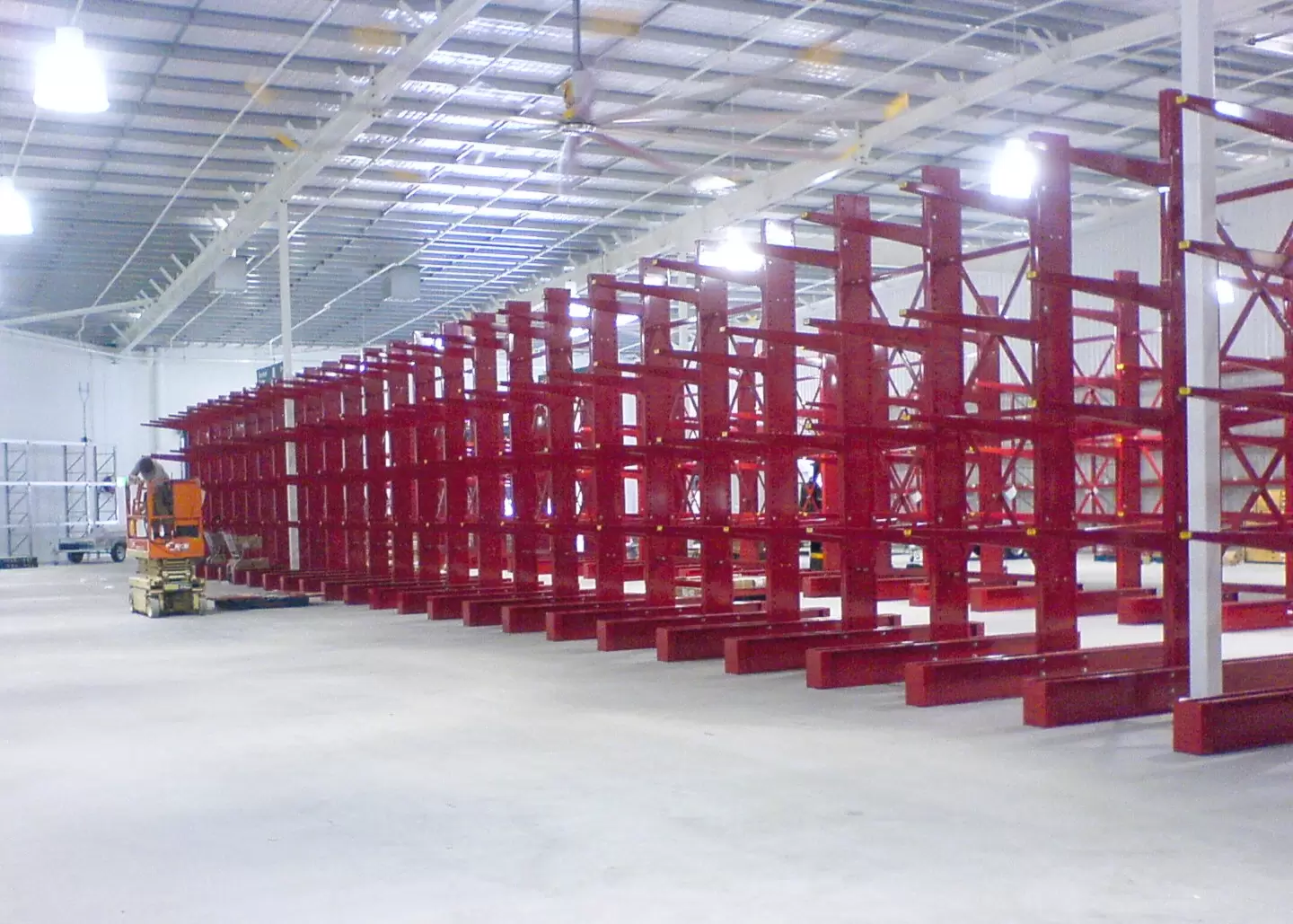 Large warehouse interior with multiple rows of red industrial storage racking extending beneath a high exposed ceiling.