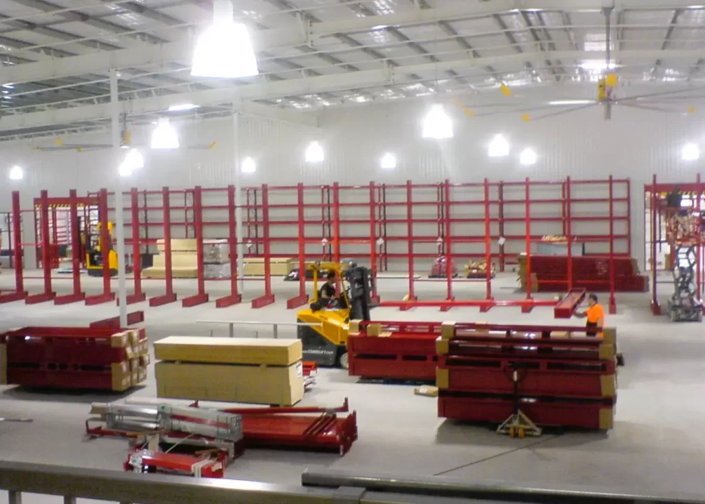 Warehouse installation area with red cantilever racking, stacked steel components, a yellow forklift, and workers beneath bright industrial lighting.