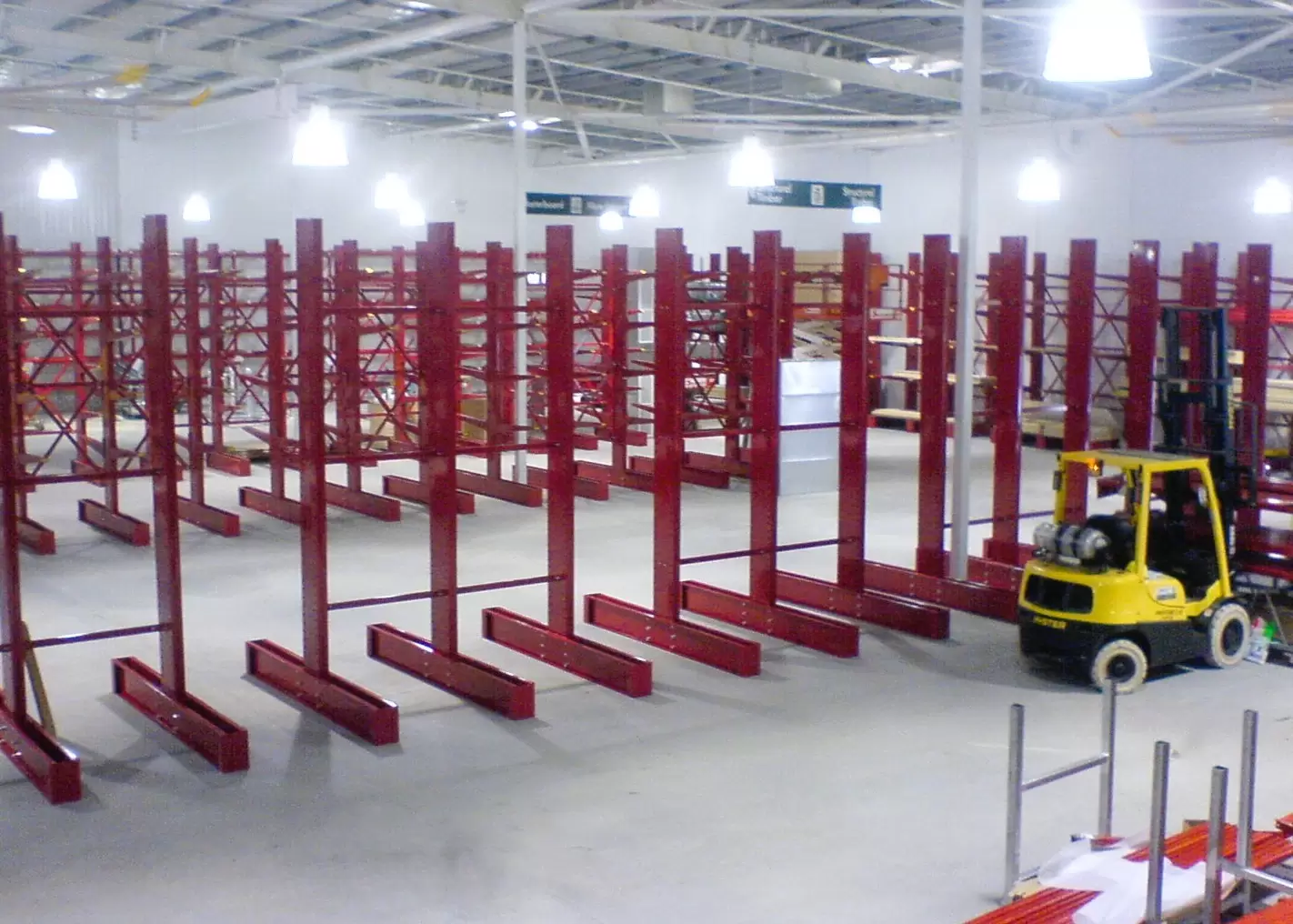 Industrial warehouse with rows of red cantilever storage racks and a yellow forklift positioned in a wide concrete aisle.