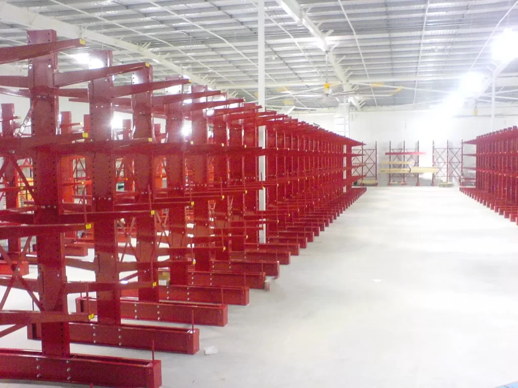 Wide warehouse aisle bordered by long rows of red cantilever storage racks beneath a high industrial ceiling with bright overhead lighting.