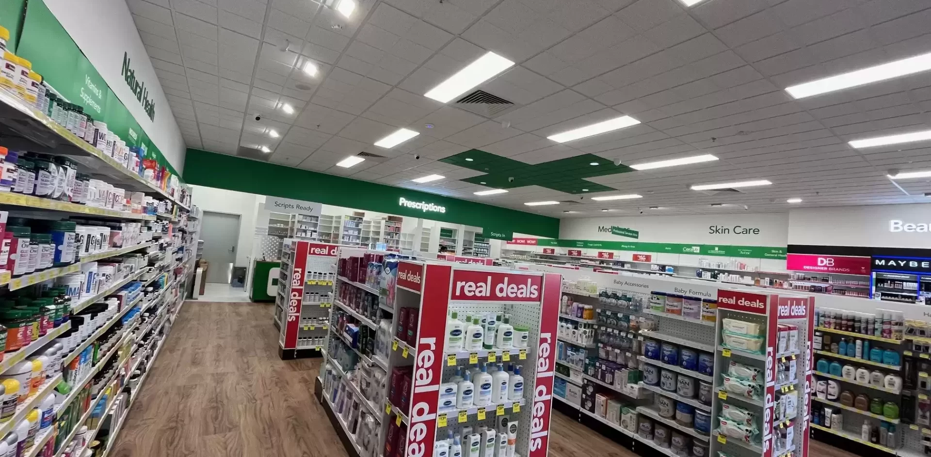 Interior of a pharmacy and beauty retail store showing product aisles, “Real Deals” promotional displays, timber-look flooring, and overhead department signage.