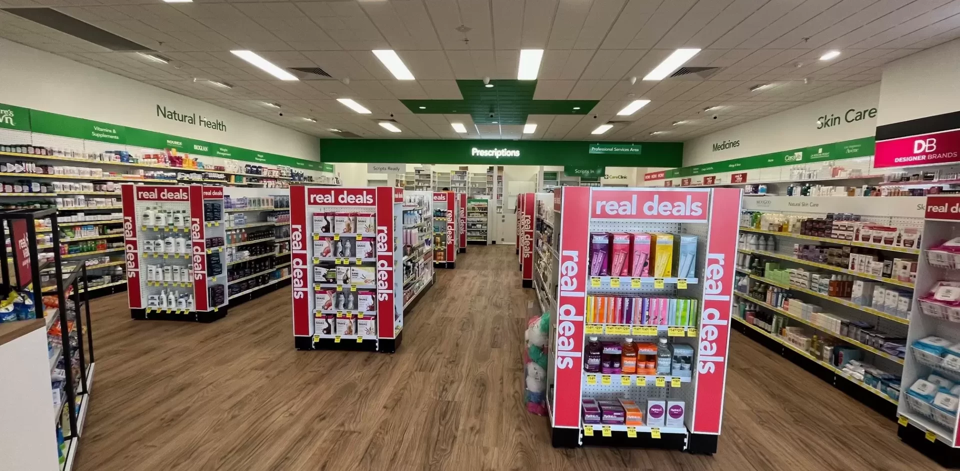 Wide view of a pharmacy retail interior with timber-look flooring, stocked product aisles, red “Real Deals” promotional displays, and green department signage.
